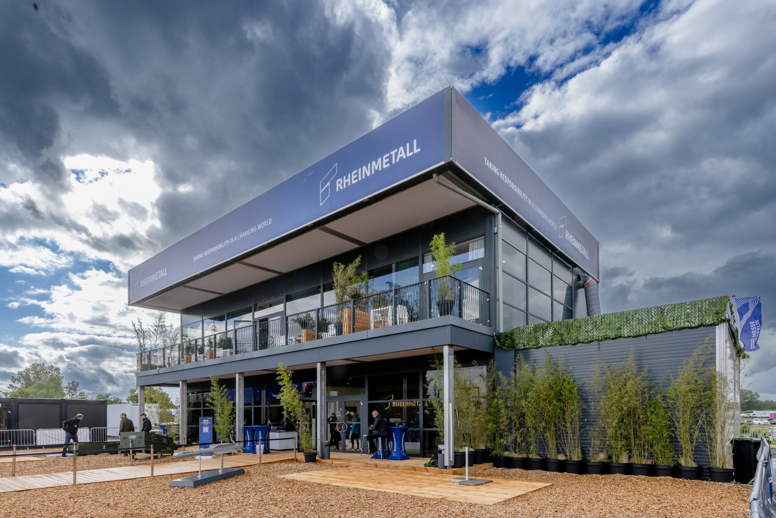 Der zweigeschossige Rheinmetall-Pavillon im Freigelände mit umlaufendem Balkon, Glasfassade und bepflanztem Vorplatz.