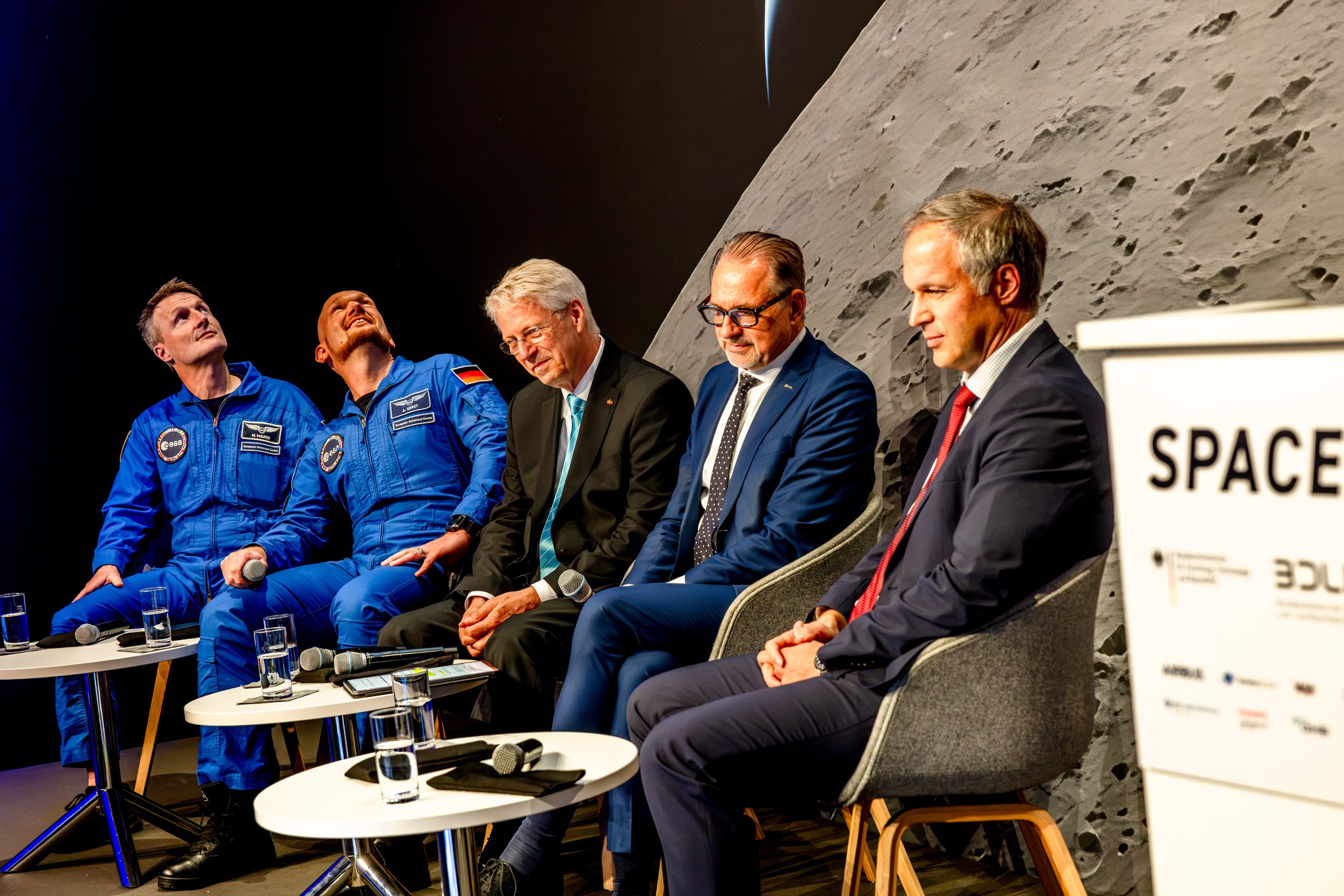 Matthias Maurer and Alexander Gerst sit in blue ESA flight suits with three further panel guests on the ILA STAGE Space 