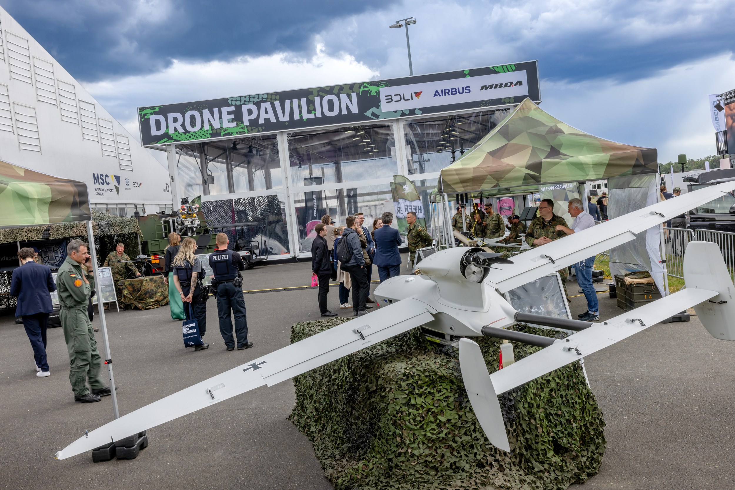 Eine große militärische Starrflügeldrohne steht auf einem mit Tarnnetz verkleideten Sockel vor dem Drone Pavilion der ILA Berlin 2026. Ein schwarzes Bundeswehrkreuz ist auf dem Flügel sichtbar; dahinter sprechen Besucher mit uniformierten Bundeswehrangehörigen, während zwei Polizeikräfte links am Stand stehen.