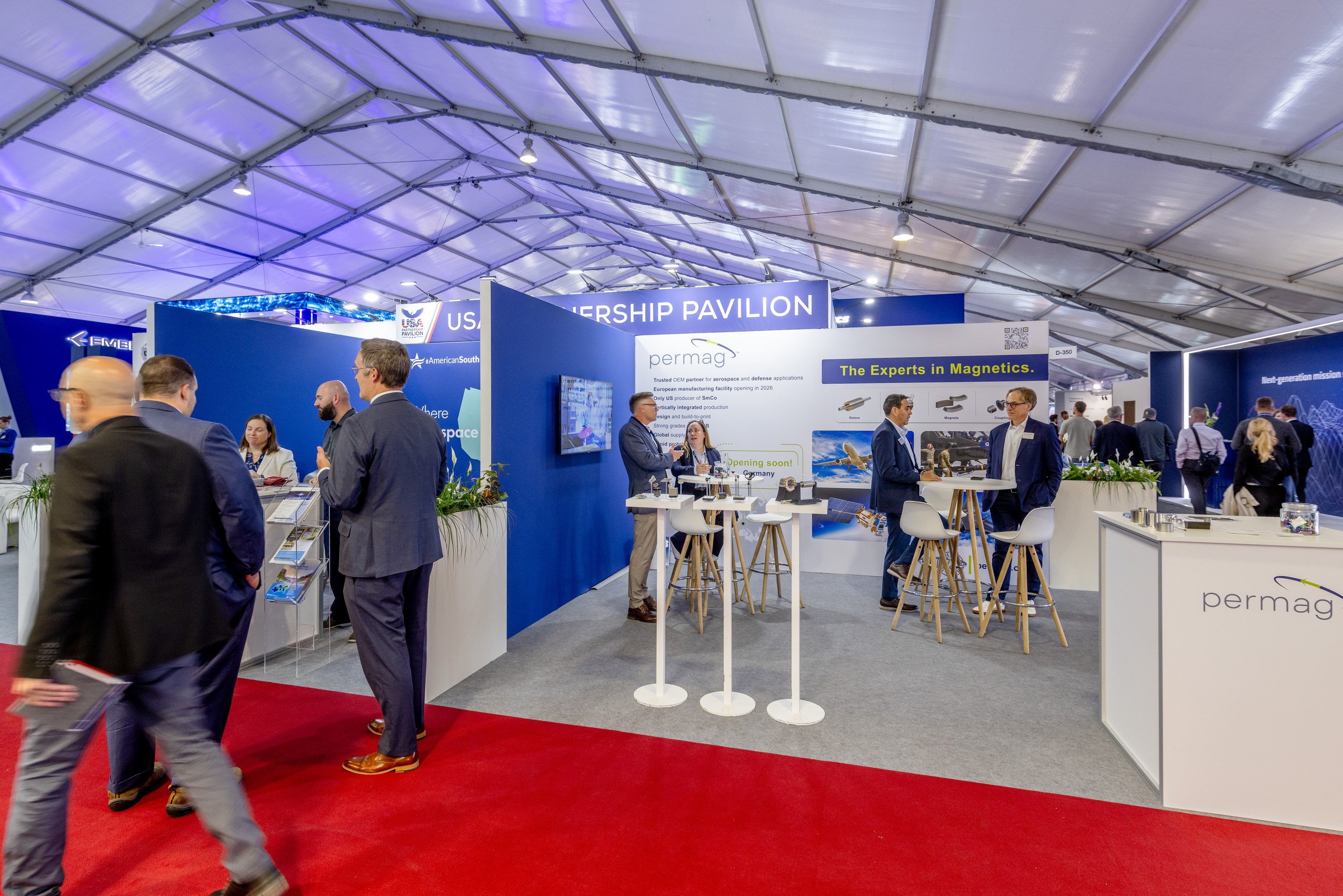 Blick in den USA Partnership Pavilion in Halle D: Fachbesucher an Hochtischen vor blau-weißen Standwänden am roten Gang.