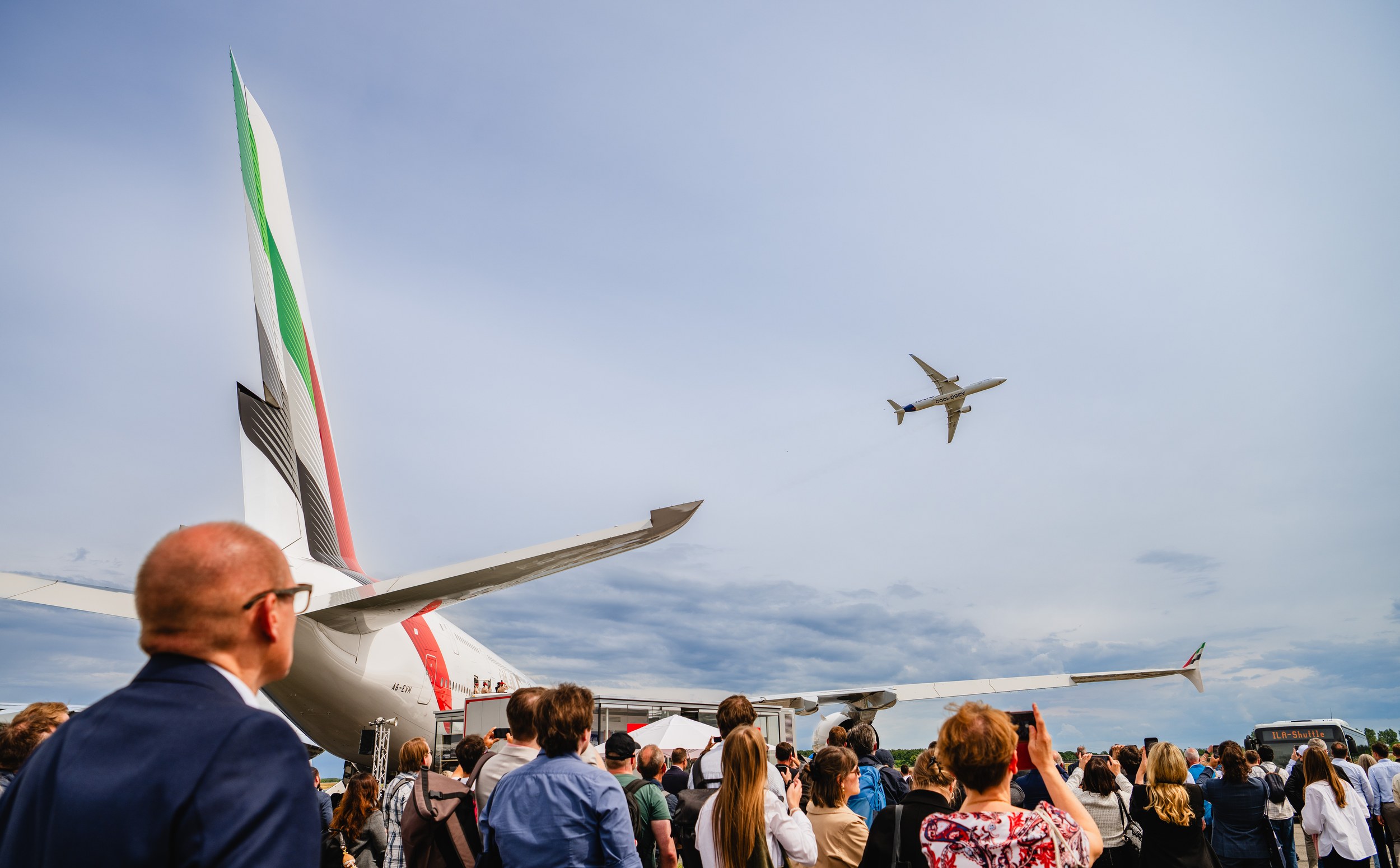 Eine große Besuchergruppe verfolgt und fotografiert einen Airbus A350 im Überflug über dem Freigelände.