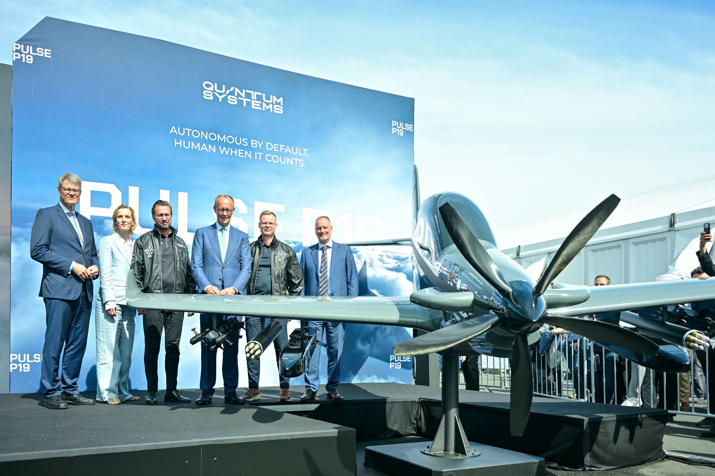 Friedrich Merz poses with five other official guests on the Quantum Systems stage before the PULSE P19. Its propeller and right wing project prominently into the foreground, multi-barrel military stores are visible beneath the wings and the branded backdrop fills the scene.