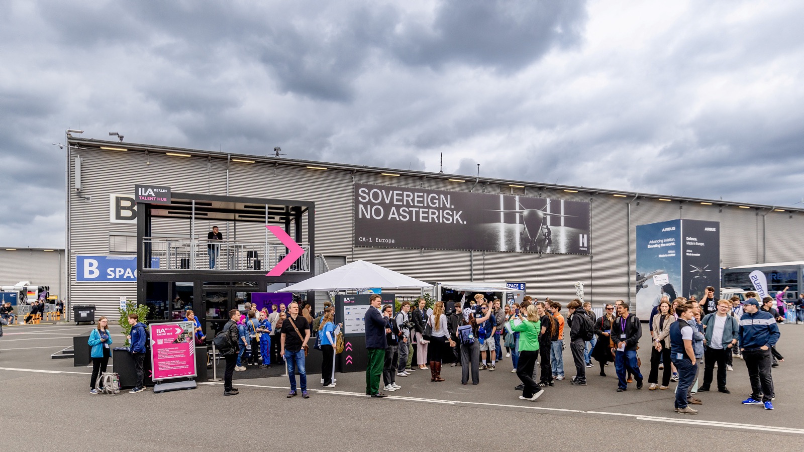 Großes Werbebanner an der Fassade eines zweigeschossigen Messebaus, davor wartende Besucher.